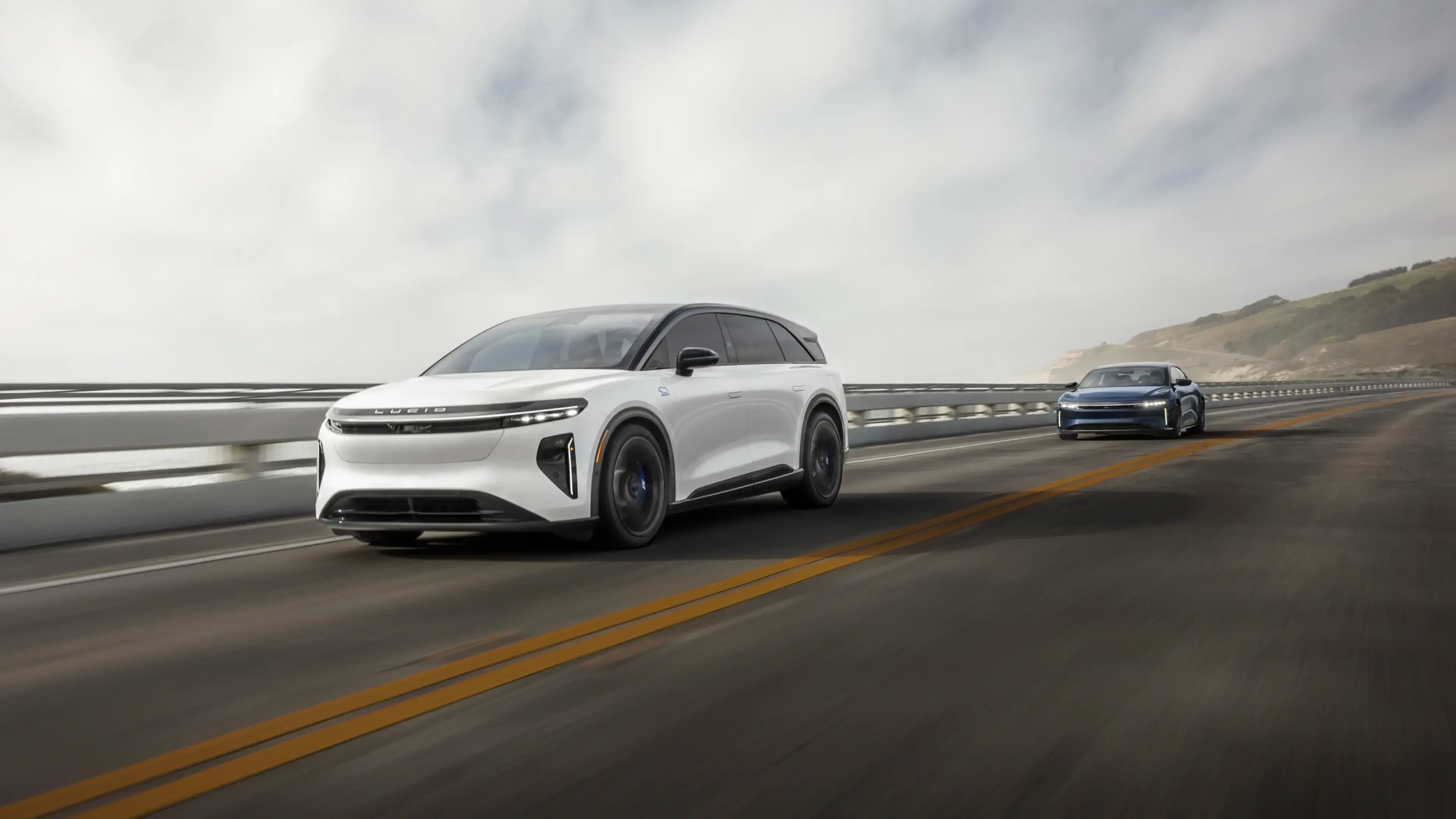 A white Lucid Gravity SUV leads a blue Lucid Air sedan on a coastal highway under a cloudy sky.