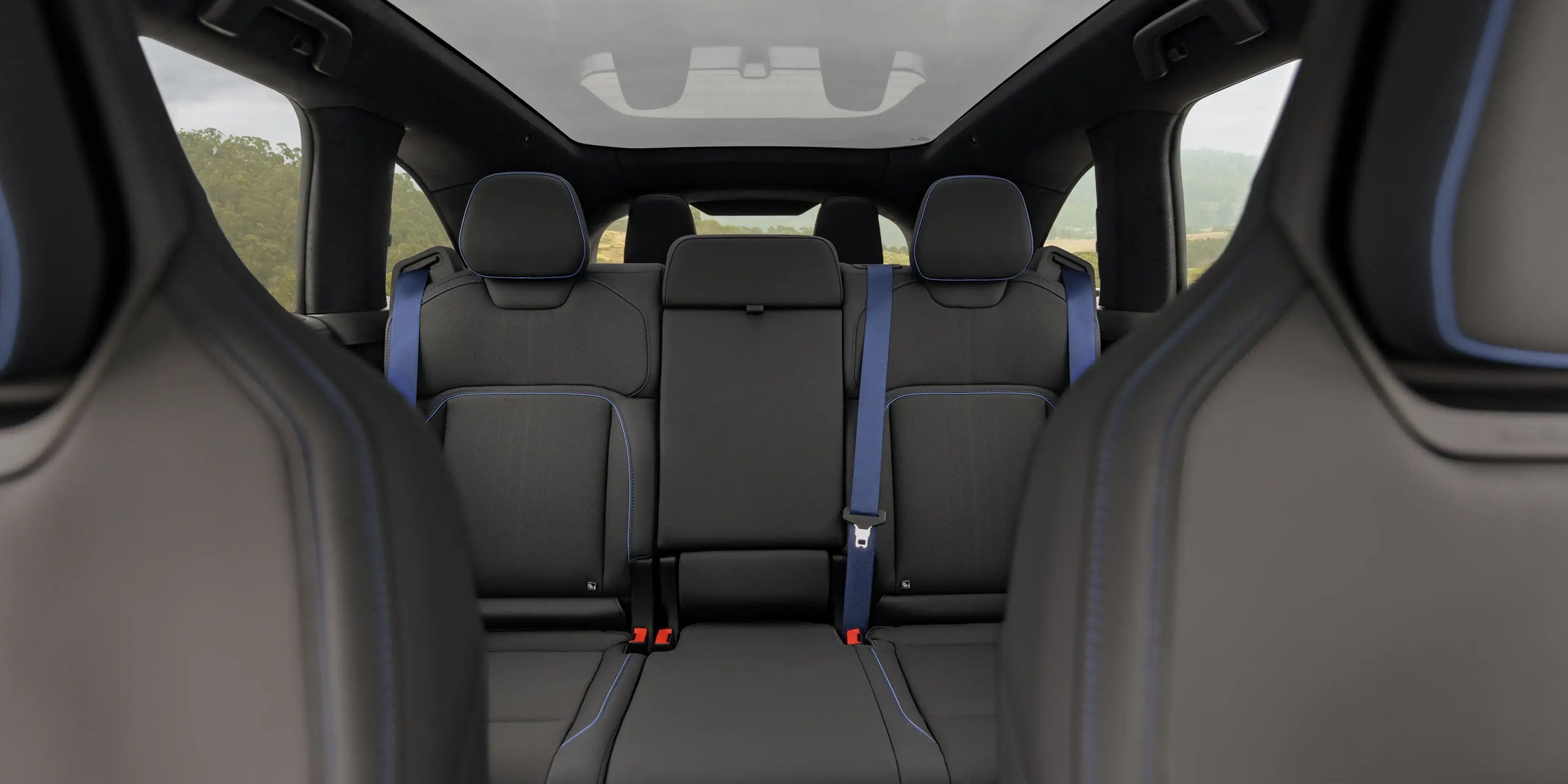 Car interior rear seats in black with blue stitching and blue seatbelts, panoramic roof, green landscape visible through windows.