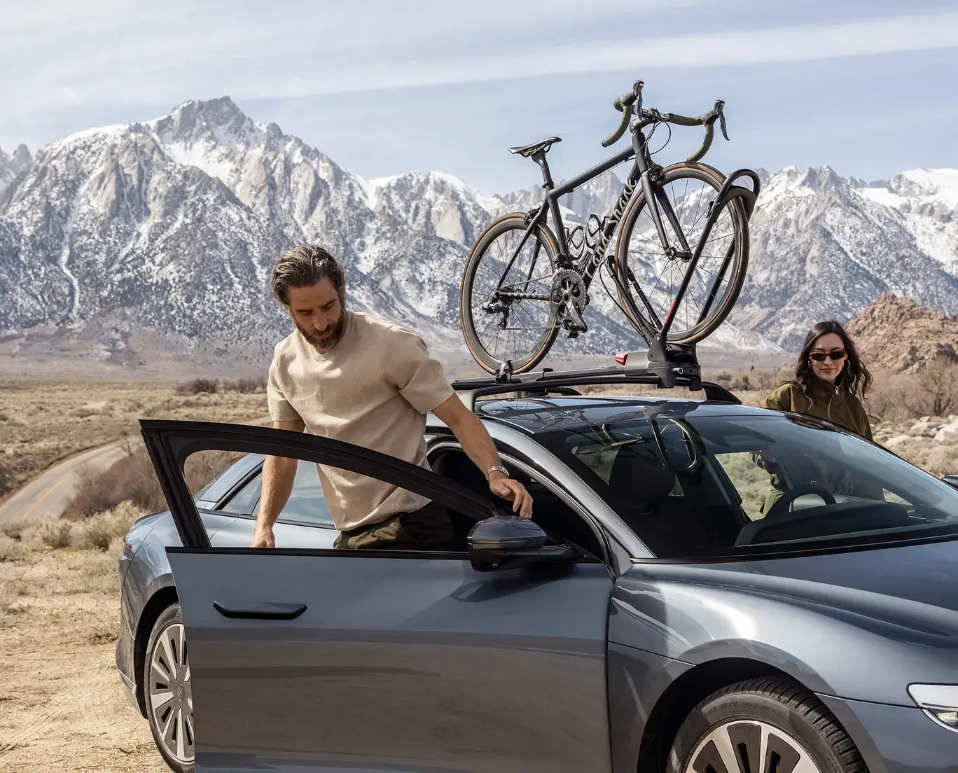 Car with roof-mounted bicycle parked in desert landscape with snow-capped mountains in background.