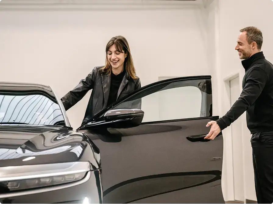 A smiling woman and man in black outfits examine a Lucid Air car with an open door in a bright showroom.