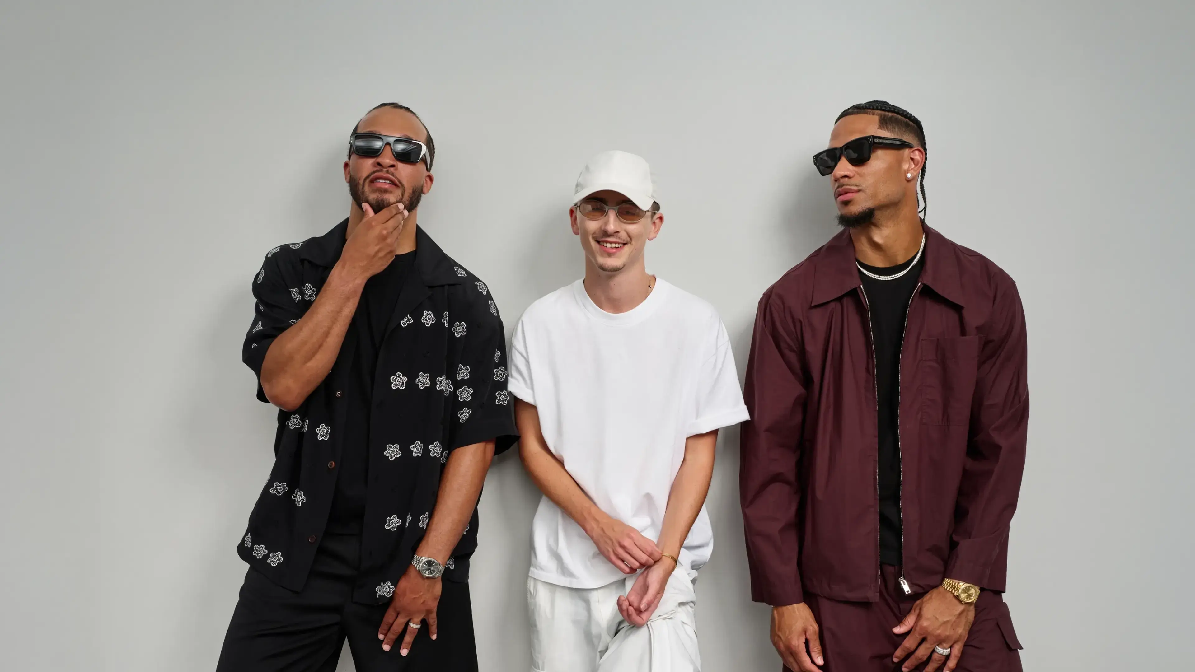 Timothée Chalamet, Josh Hart, and Jalen Brunson posing against gray wall - Jalen in black patterned shirt, Timothée in white outfit, Josh in burgundy jacket.