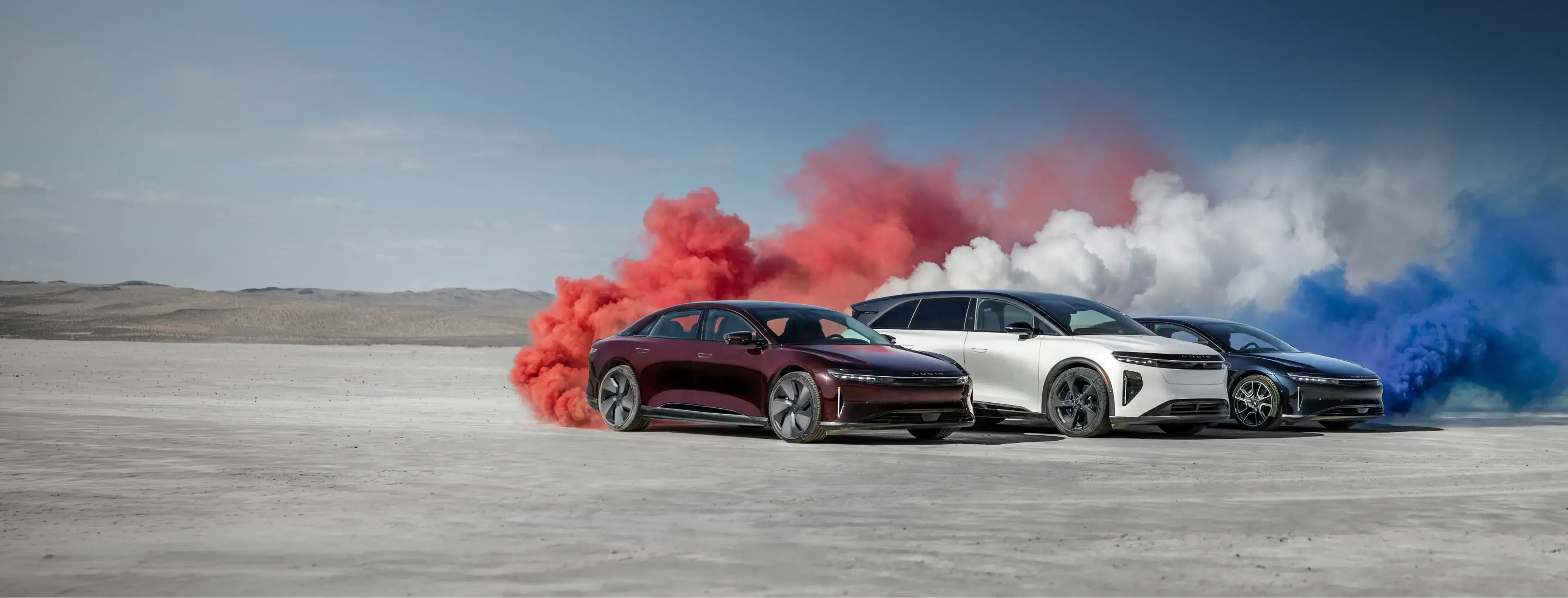 Three Lucid Air electric cars in red, white, and blue on a desert flat with colorful smoke clouds behind each.