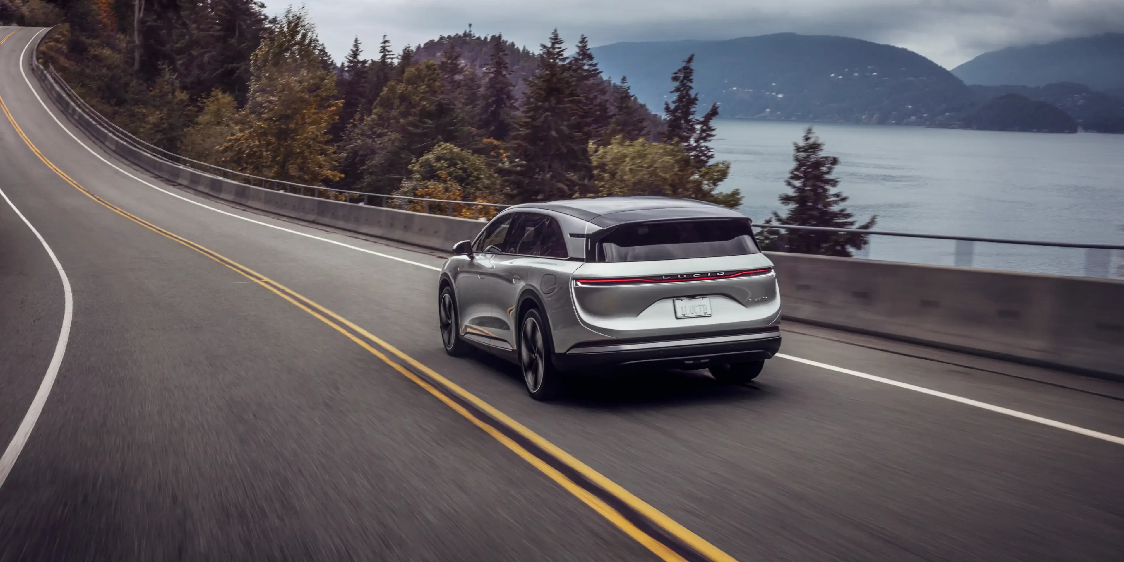 Lucid Gravity driving on coastal highway with evergreen trees and mountains overlooking misty water.