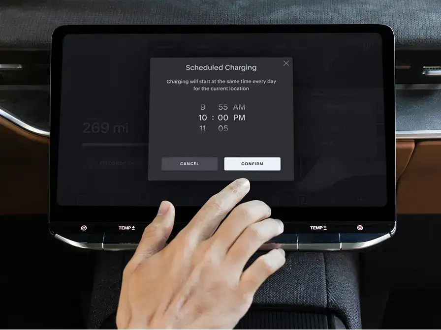 A hand reaching toward a car touchscreen displaying a "Scheduled Charging" dialog set to 10:00 PM.
