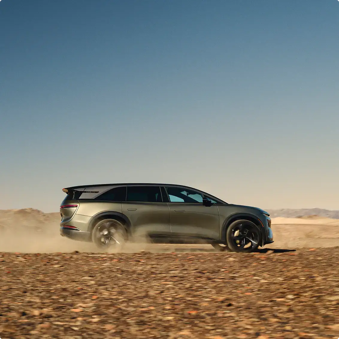 A silver-green SUV drives through a dusty desert landscape, kicking up dust under a clear blue sky.