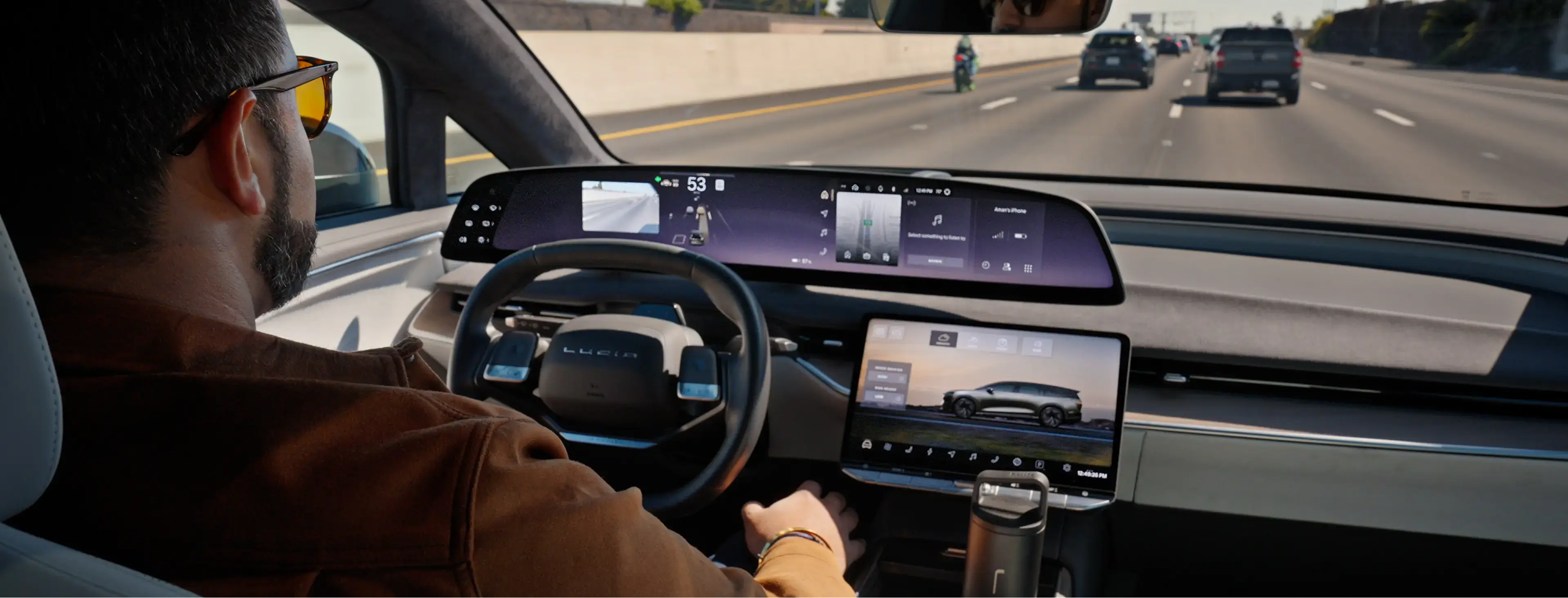 Interior view of a Lucid electric vehicle with dual touchscreens, driver's hands on wheel, highway visible ahead.