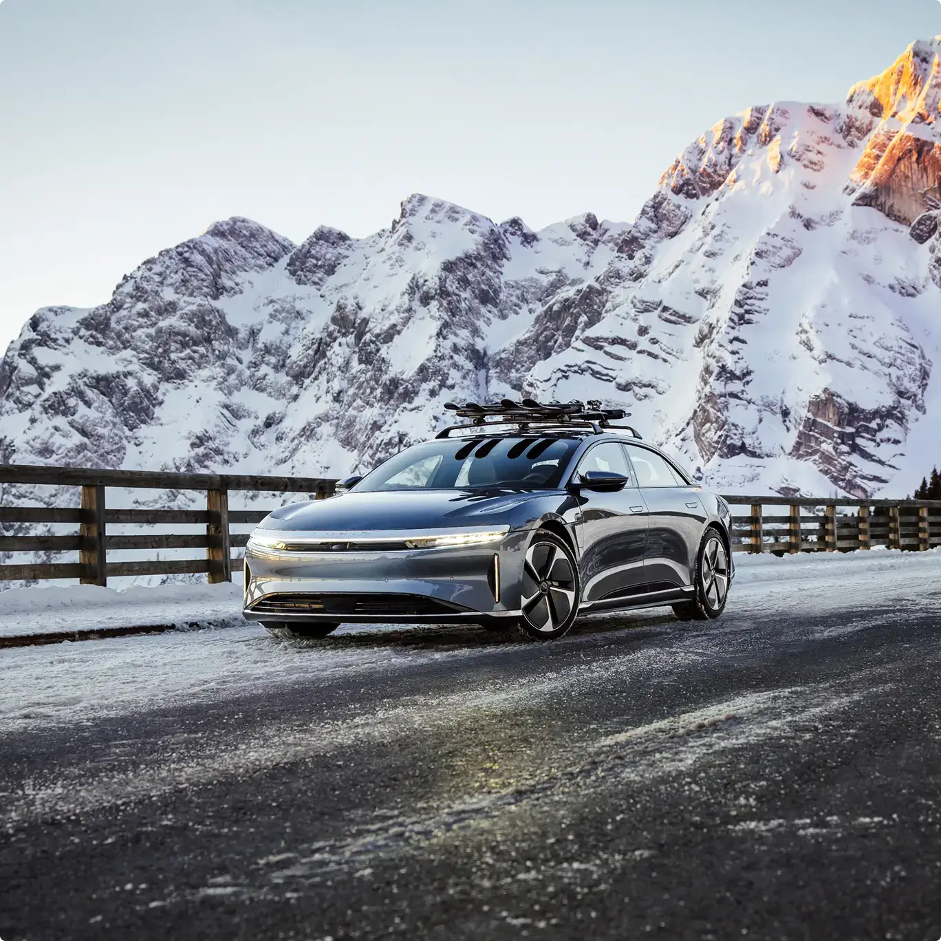 Sleek silver electric car with roof rack parked on snowy mountain road against backdrop of snow-covered peaks.