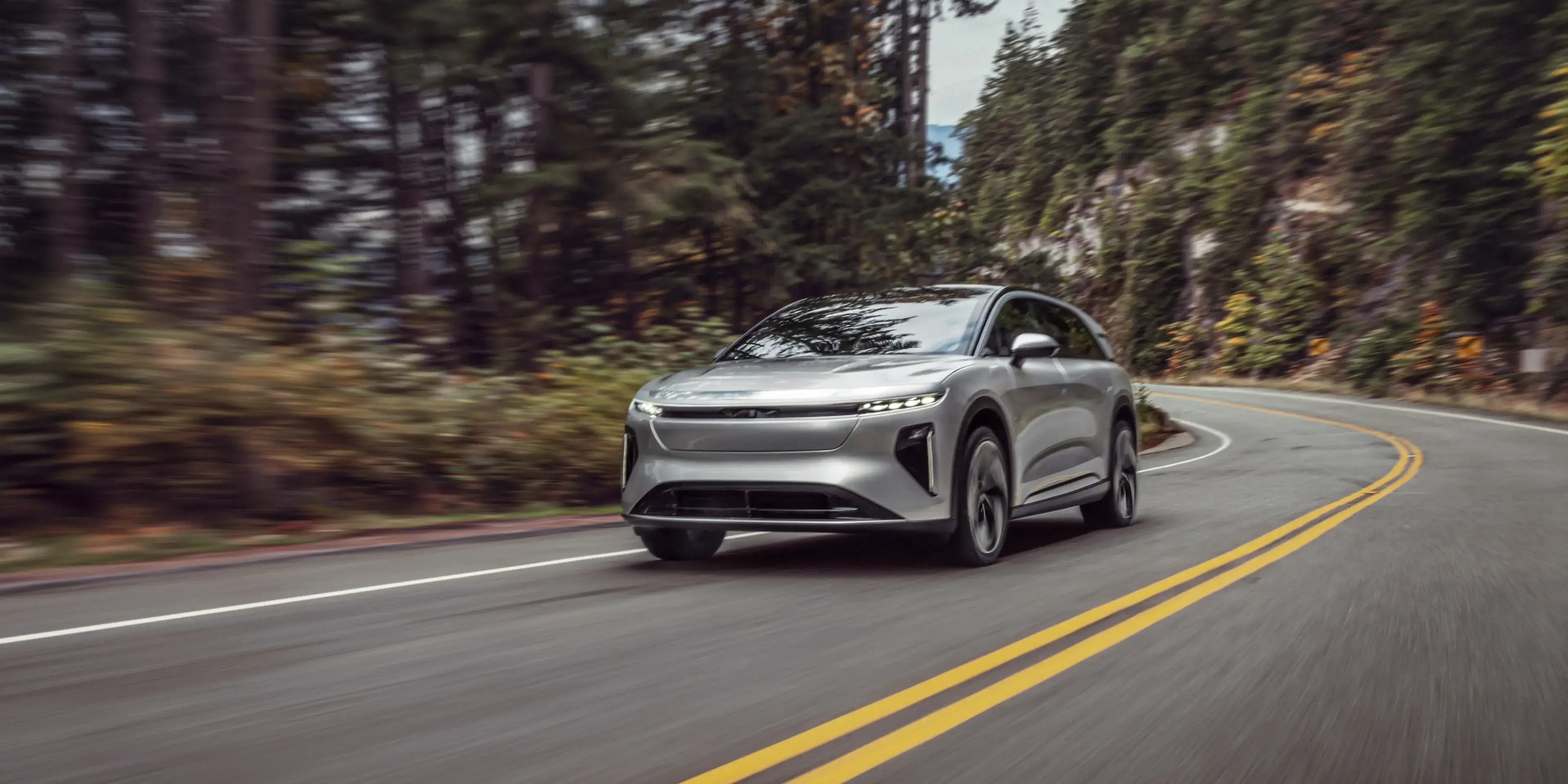 Une Lucid Gravity roulant sur une route forestière sinueuse avec de grands conifères et de l’eau visible au loin.