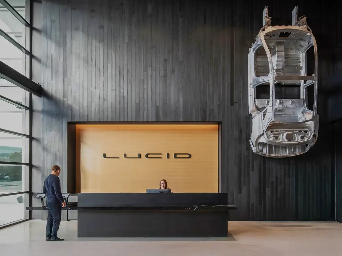 Lucid Motors reception area with sleek black desk, gold-lit logo wall, and car chassis displayed as wall art.