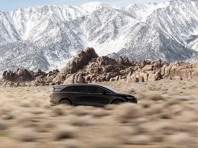 Black SUV driving through a desert landscape with rocky terrain and snow-capped mountains in the background.