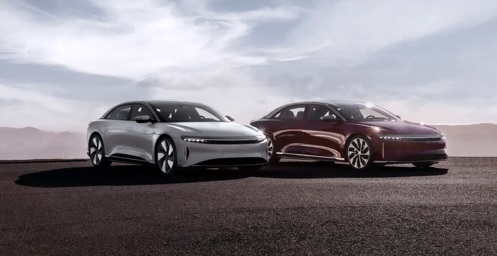 Two sleek electric sedans, one silver and one burgundy, parked side by side on dark ground under a cloudy sky.