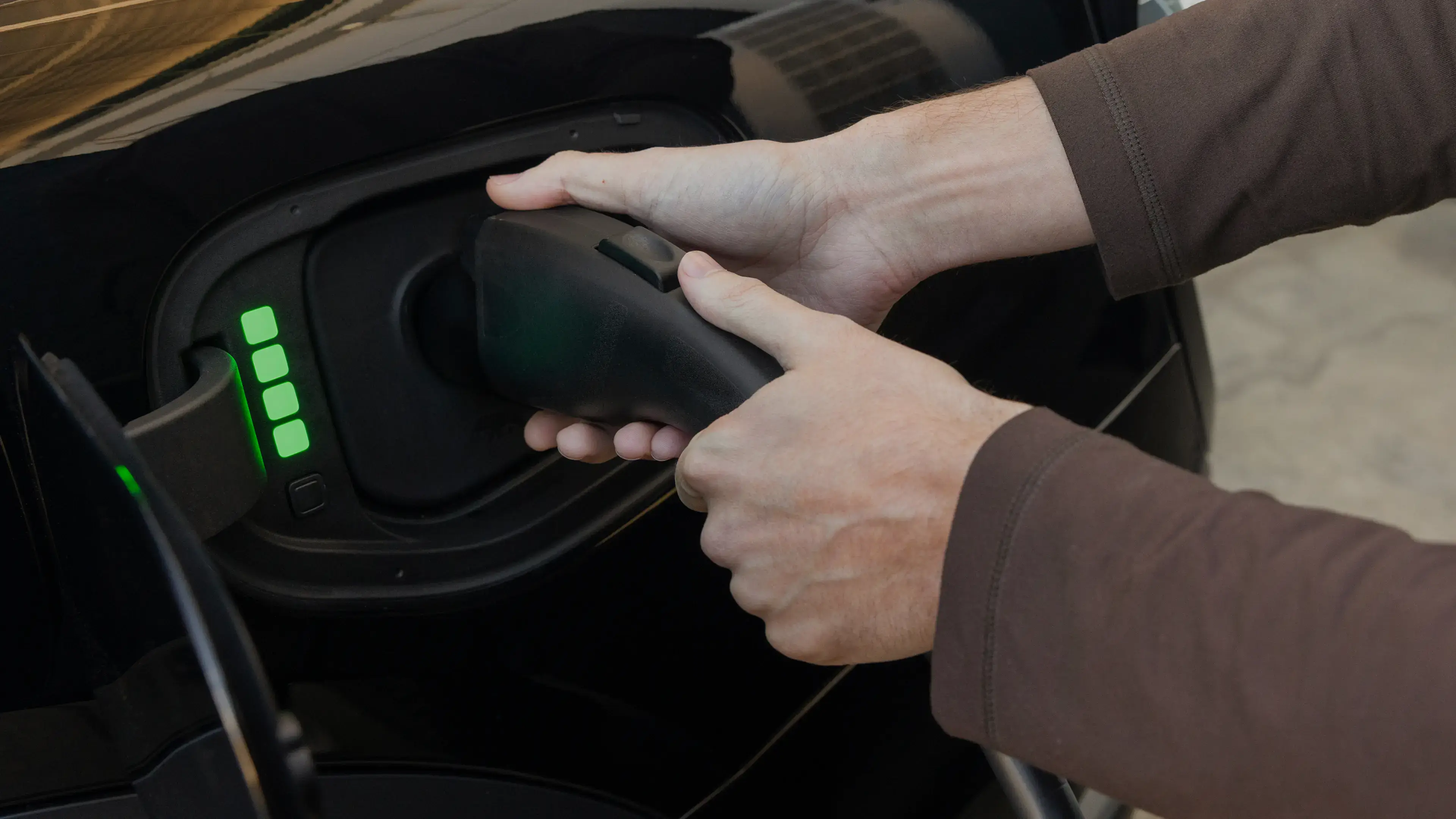 Hands plugging in an electric vehicle charging cable with green battery indicator lights showing charge level.