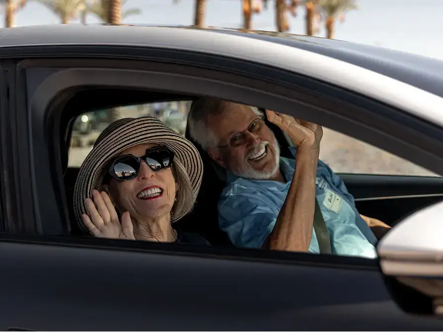 Two smiling seniors wave from a car window; woman wears a sun hat and sunglasses, man in a blue shirt beside her.
