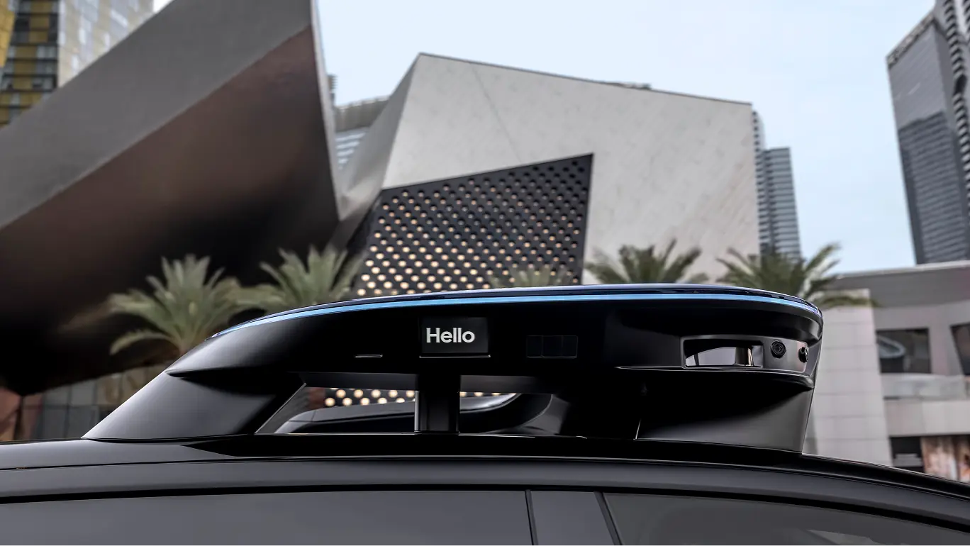 Schwarze Fahrzeugsensor-Einheit zeigt „Hello“ vor modernem Architekturgebäude mit Palmen und Stadtsilhouette.