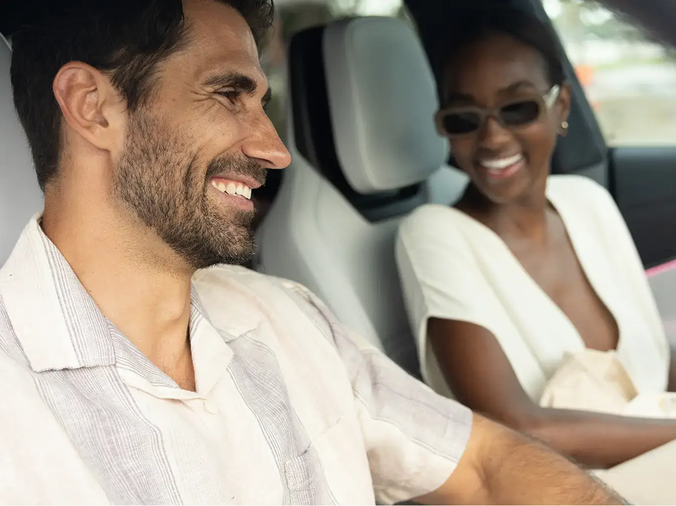 Two smiling passengers in white outfits sharing a joyful moment inside a car with light-colored interior.