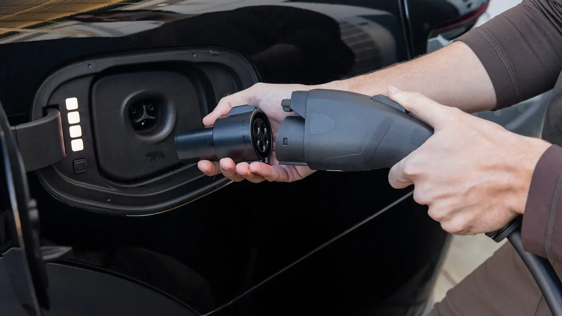 Hands connecting an electric vehicle charging plug to a black car's charging port with illuminated indicator lights.