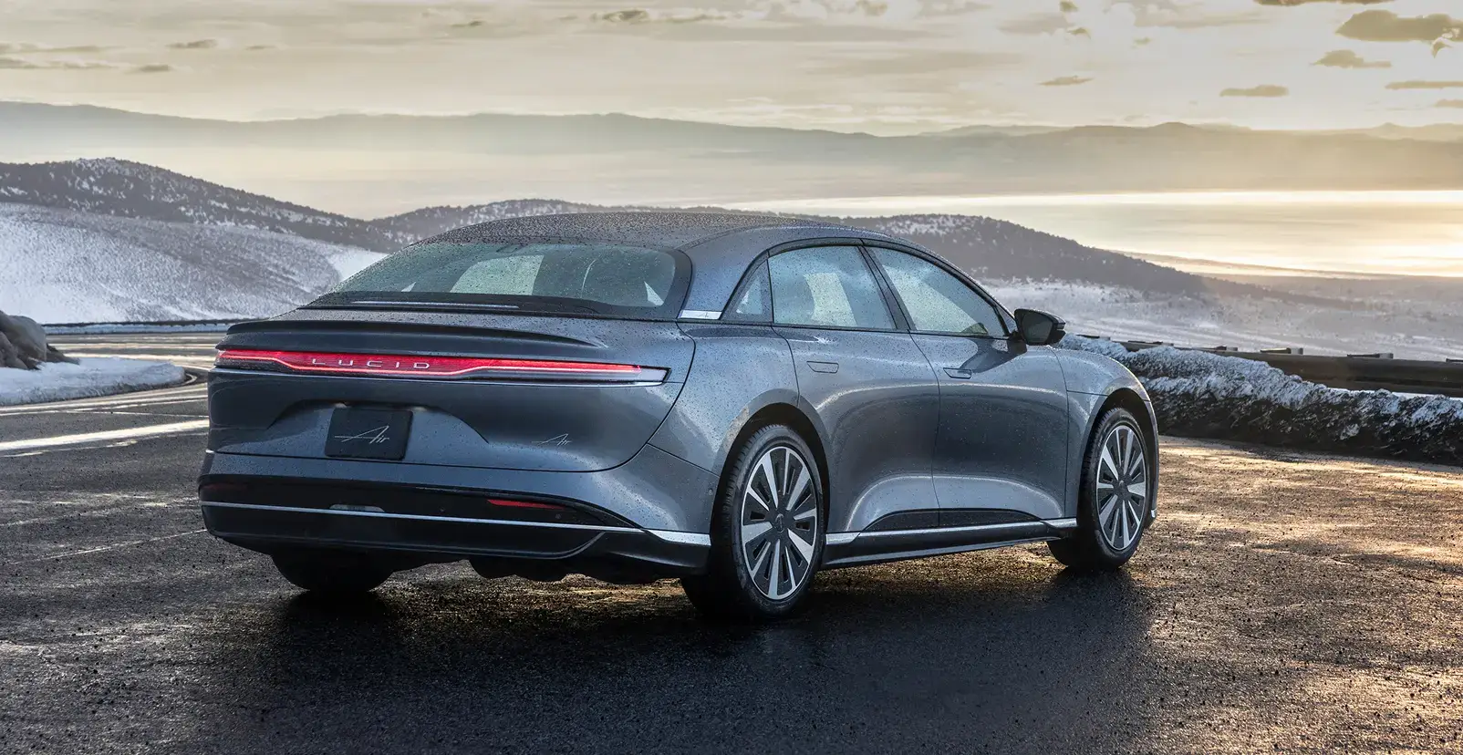 Lucid Air stationnée à un belvédère montagneux au coucher du soleil, avec un ciel nuageux spectaculaire et des collines lointaines.