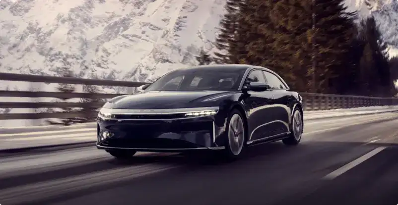 Lucid Air sedan driving on a mountain road with snow-covered peaks and evergreen trees in the background.