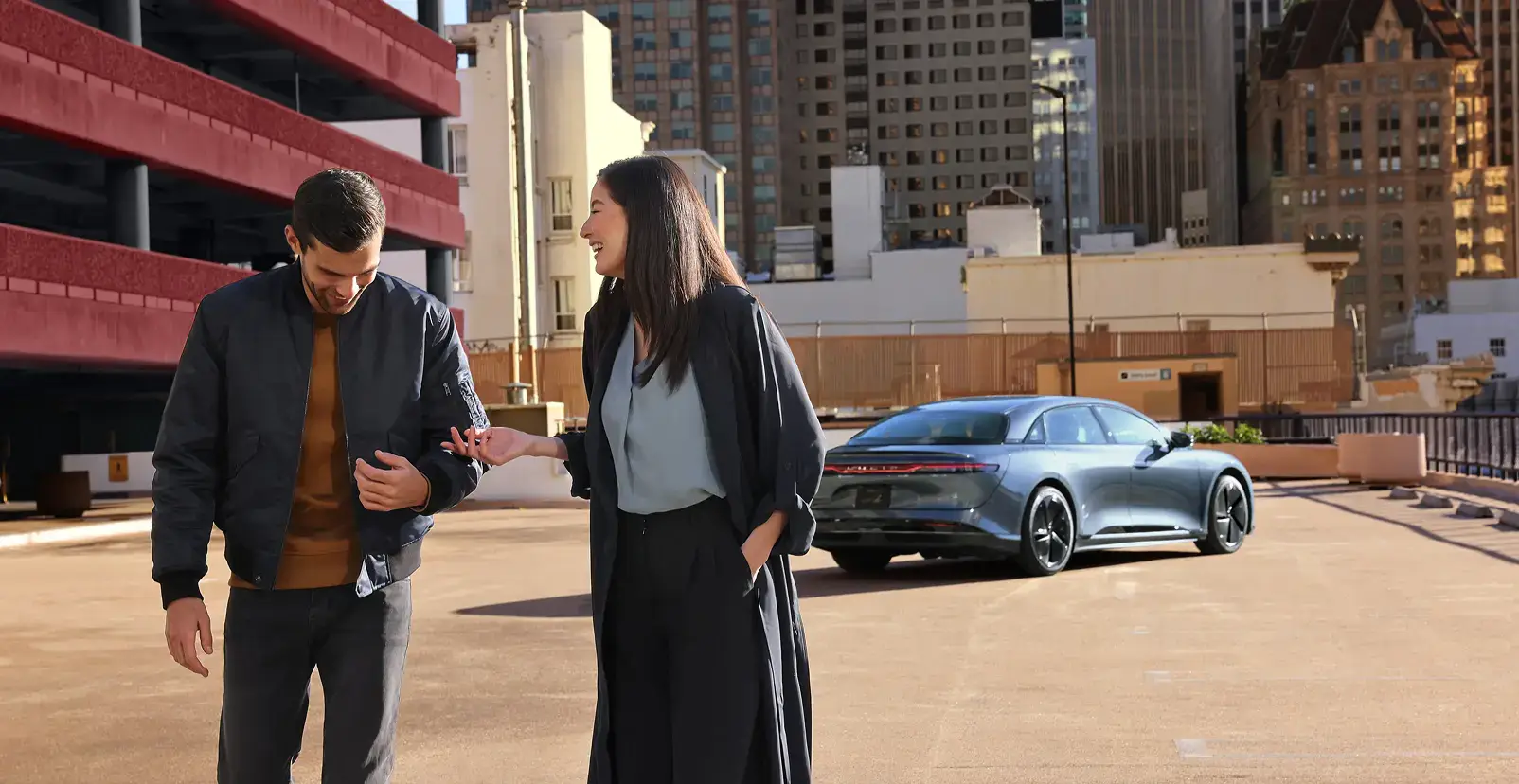 Two people walking and laughing on a rooftop parking lot with a sleek gray sedan and city skyline in the background.