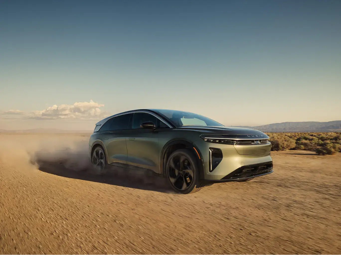 Sleek Lucid electric SUV driving through desert terrain, kicking up dust clouds against clear blue sky and distant mountains.