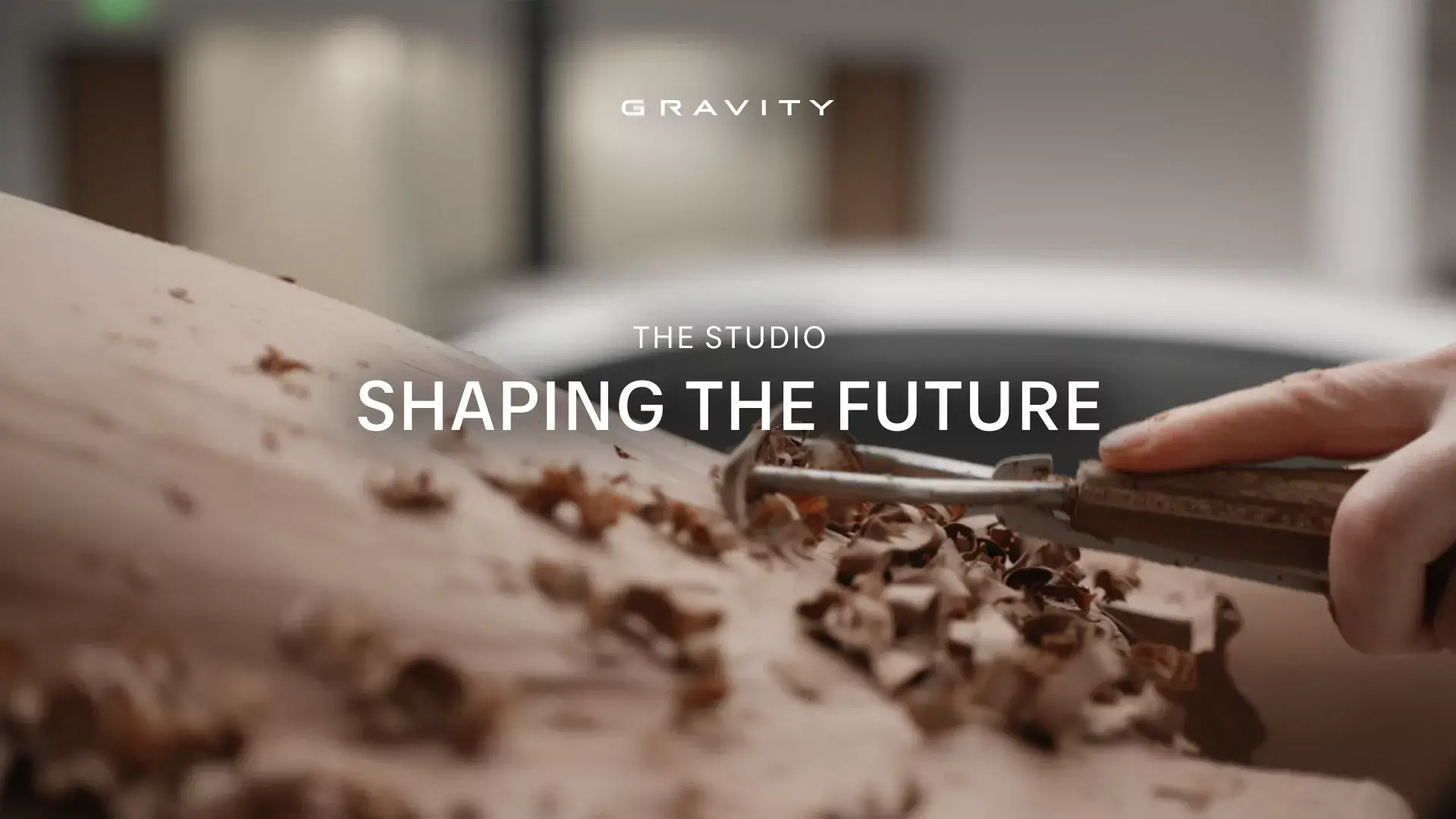 Woodworking tool carving wood with text overlay "GRAVITY THE STUDIO SHAPING THE FUTURE" in a modern workshop setting.