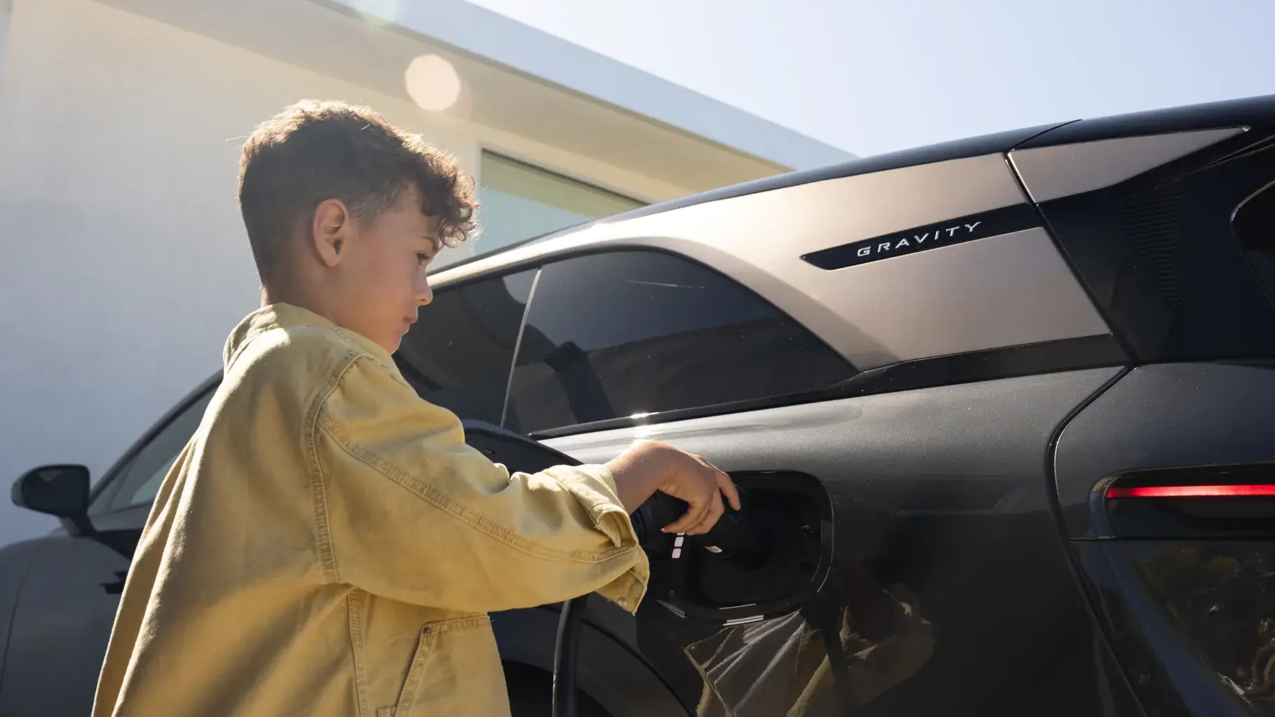 A young boy in a yellow jacket plugs in a charger on a silver Lucid Gravity electric SUV outdoors.