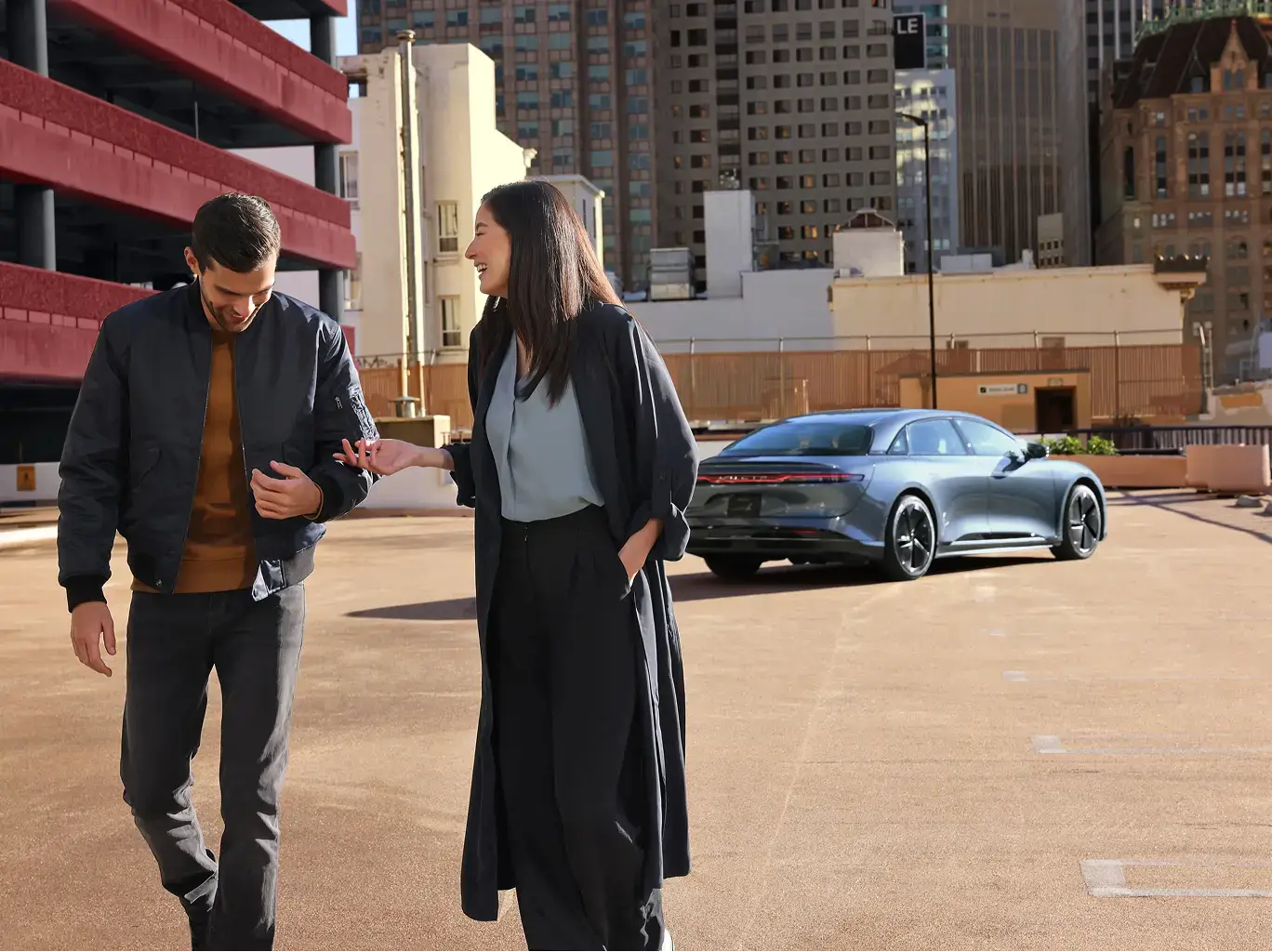 Two people conversing in an urban setting with a sleek gray Porsche electric vehicle parked behind them and city buildings.