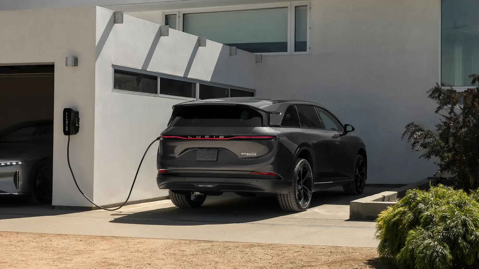 Black Lucid Gravity SUV charging at a home wall charger outside a modern white house.