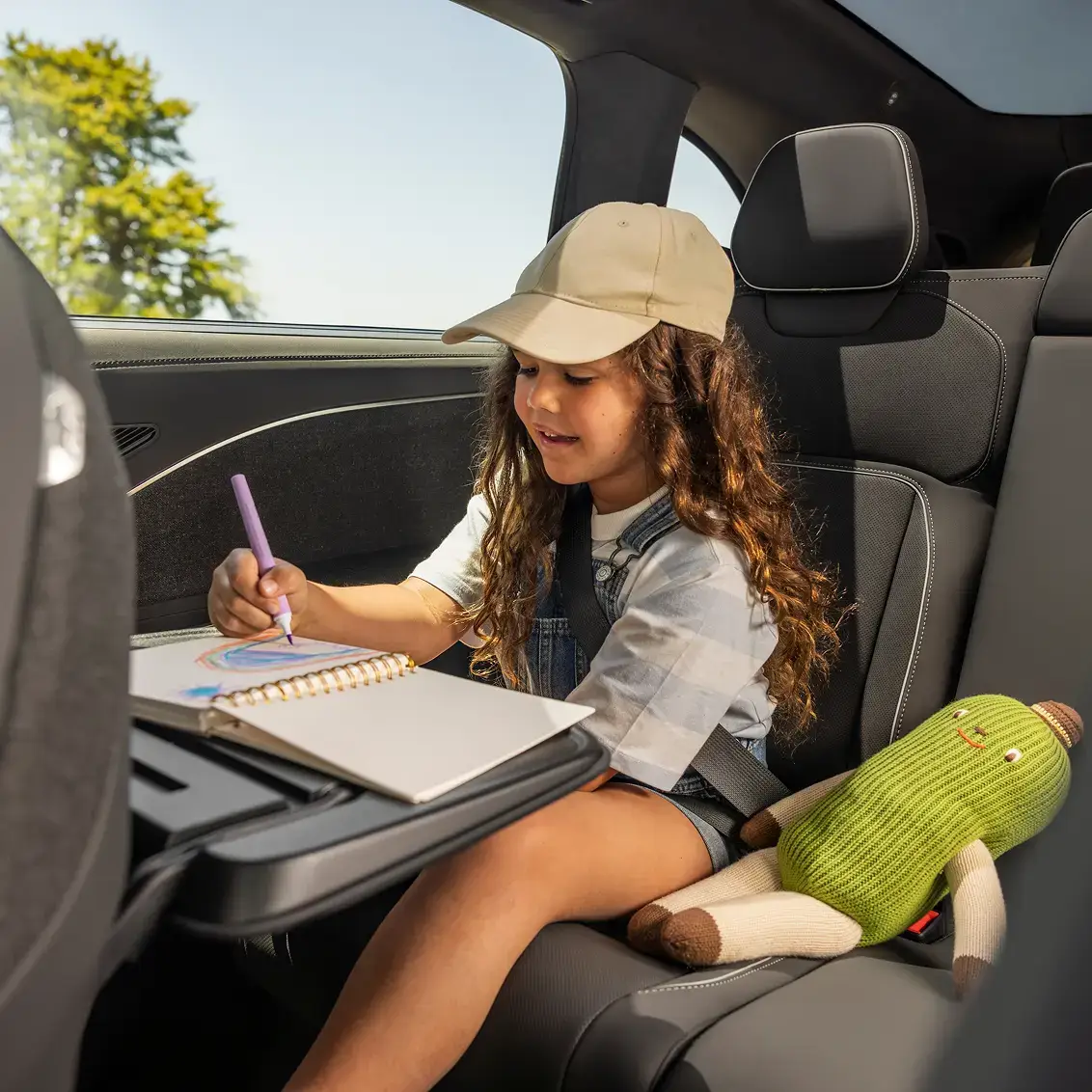 A young girl in a beige cap draws in a sketchbook in a car's back seat, with a green stuffed toy beside her.