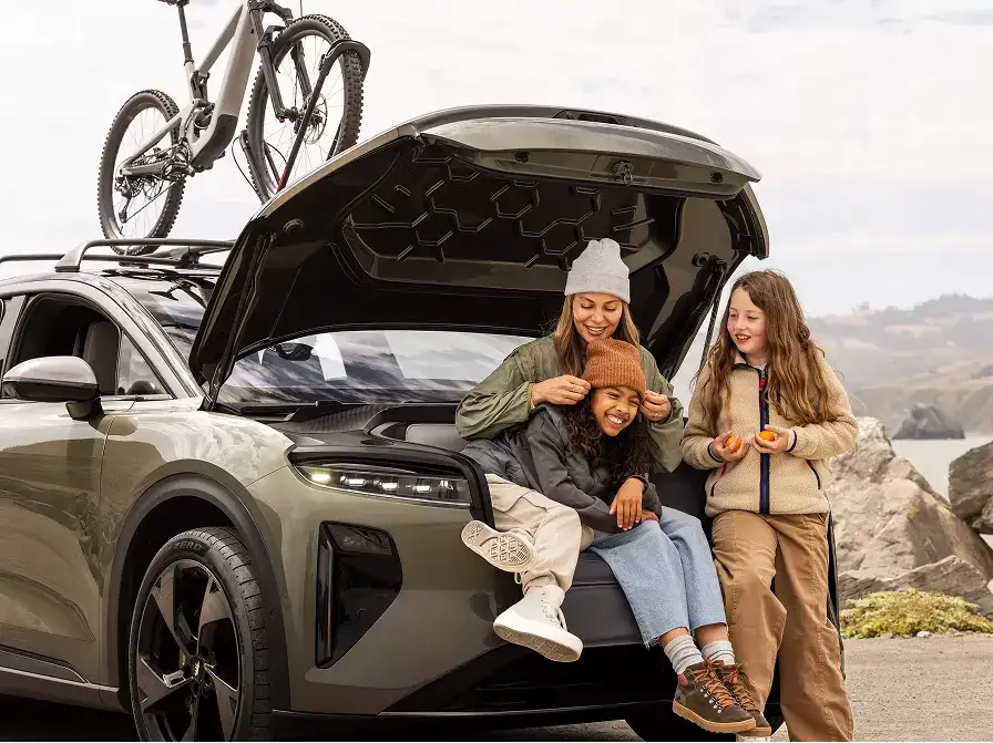 A woman and two children sit and smile at the open trunk of a Lucid Gravity SUV with a bike on the roof rack near a coastal scene.
