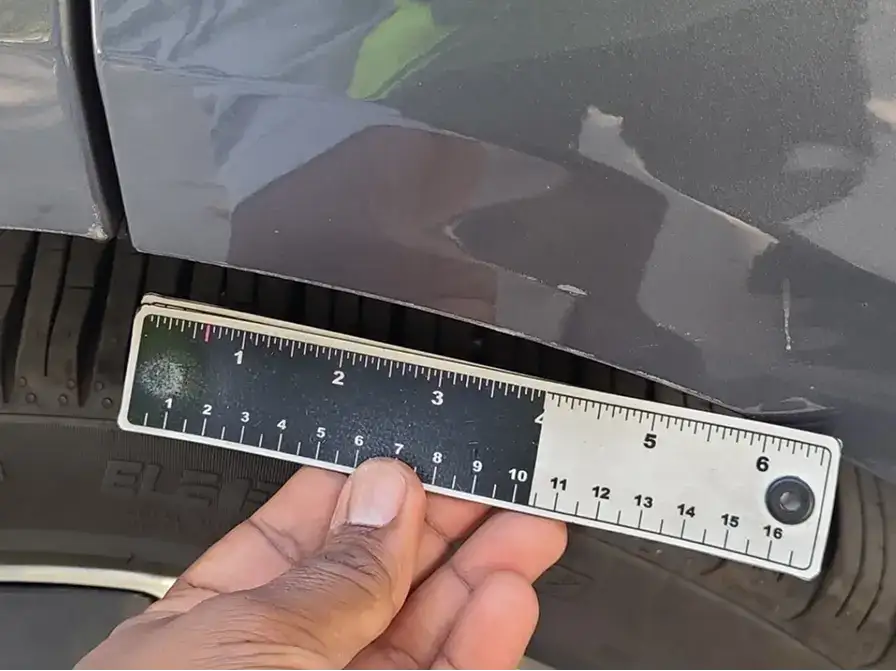 Hand holding a ruler measuring damage or gap on a dark car door panel near the tire.