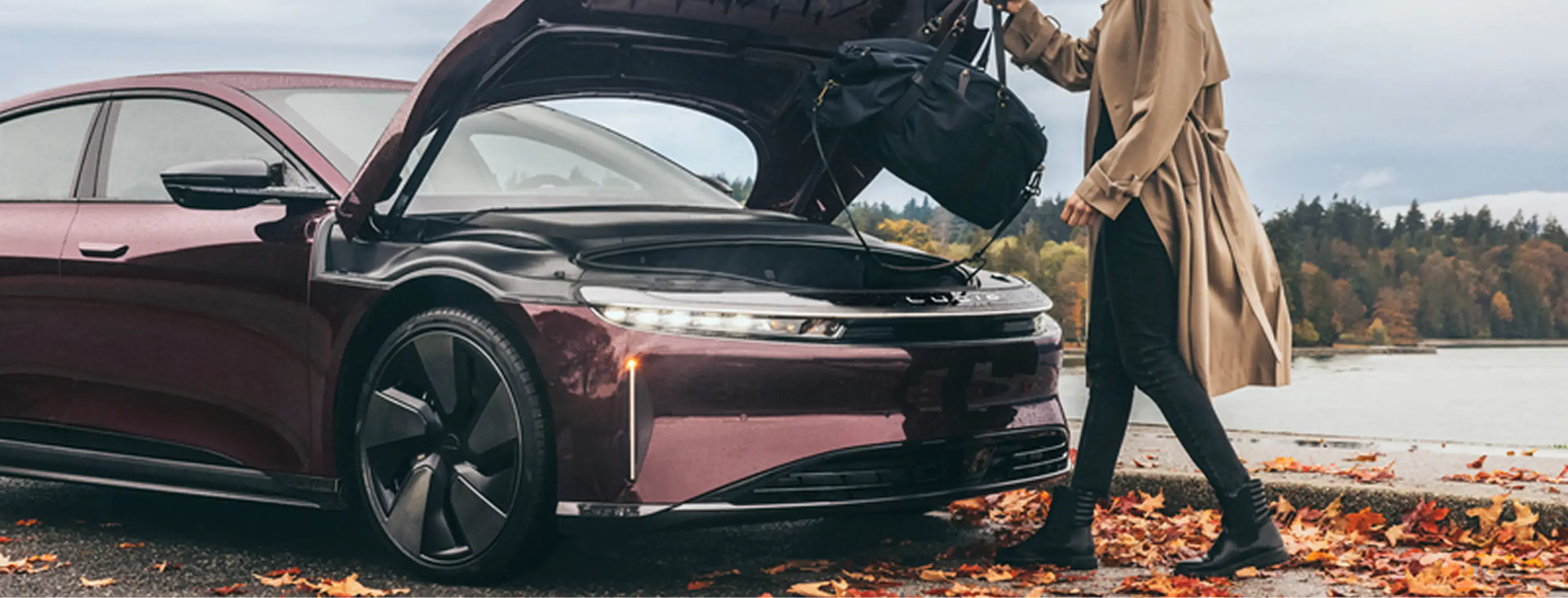 Woman in a beige coat loading a bag into the frunk of a dark red Lucid Air EV, surrounded by autumn leaves.