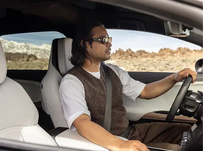Man with sunglasses and long hair driving a car with white interior, desert rocky landscape visible through the window.