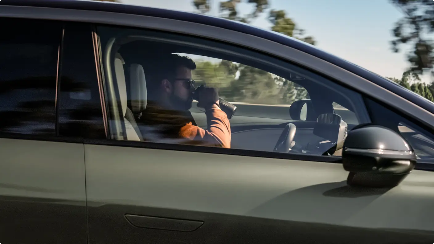 A man in an orange jacket drinks from a travel mug while sitting in the passenger seat of a green car in motion.