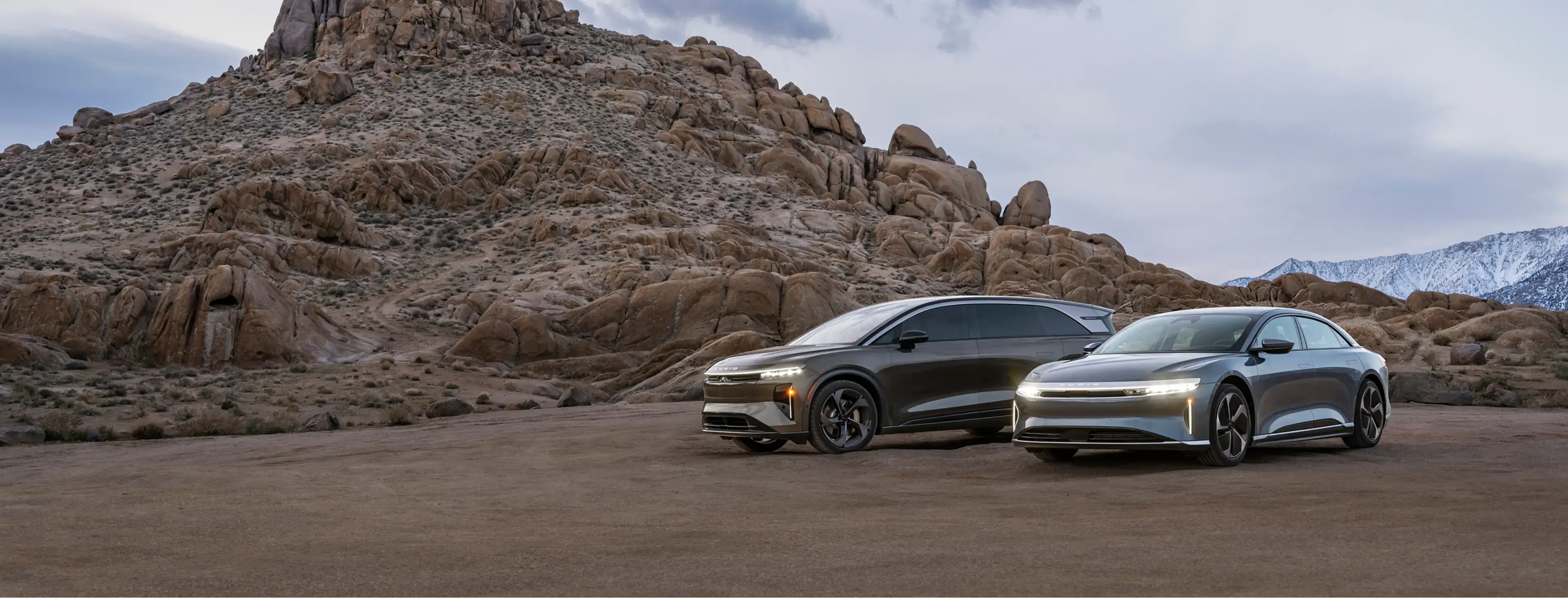 Two sleek electric vehicles with illuminated headlights parked in a desert landscape with rocky mountains in the background.