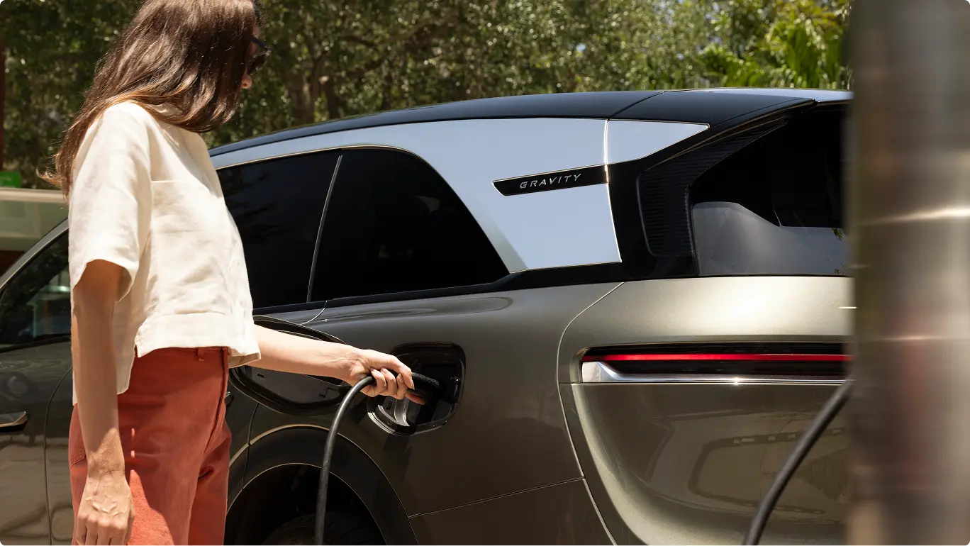 Person in white top and orange pants charging a sleek Lucid Gravity electric vehicle outdoors among trees.