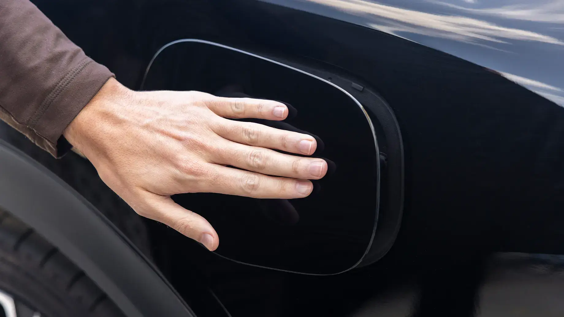 A hand pressing on a black electric vehicle charging port cover on a dark-colored car exterior.