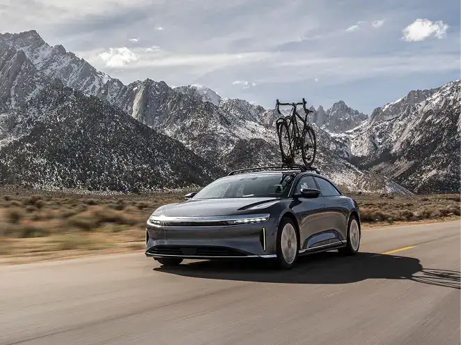 A sleek gray electric car with a bicycle mounted on the roof rack drives along a mountain highway.