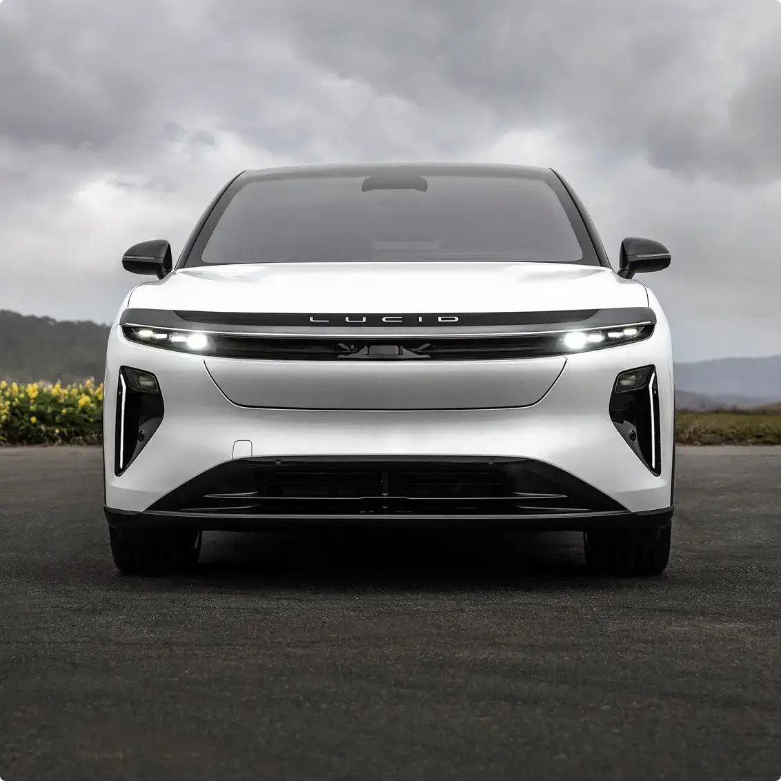 Front view of a white Lucid electric SUV with illuminated headlights parked on pavement under a cloudy sky.
