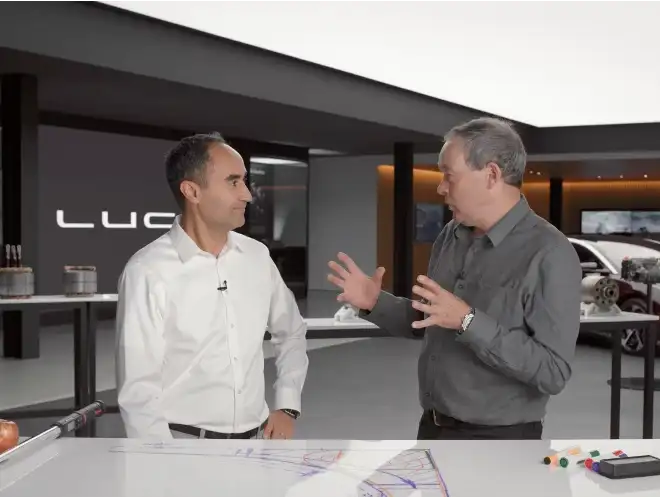 Two men in conversation at a Lucid Motors showroom, with design sketches visible on a table in the foreground.