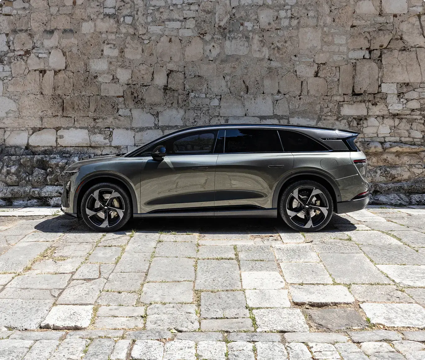 Side profile of a sleek gray SUV parked on a cobblestone surface against a rustic stone wall.