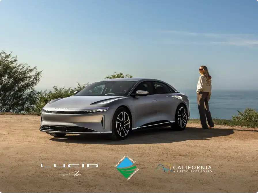 Silver Lucid Air electric sedan parked outdoors near the ocean, with a woman standing beside it under a clear blue sky.