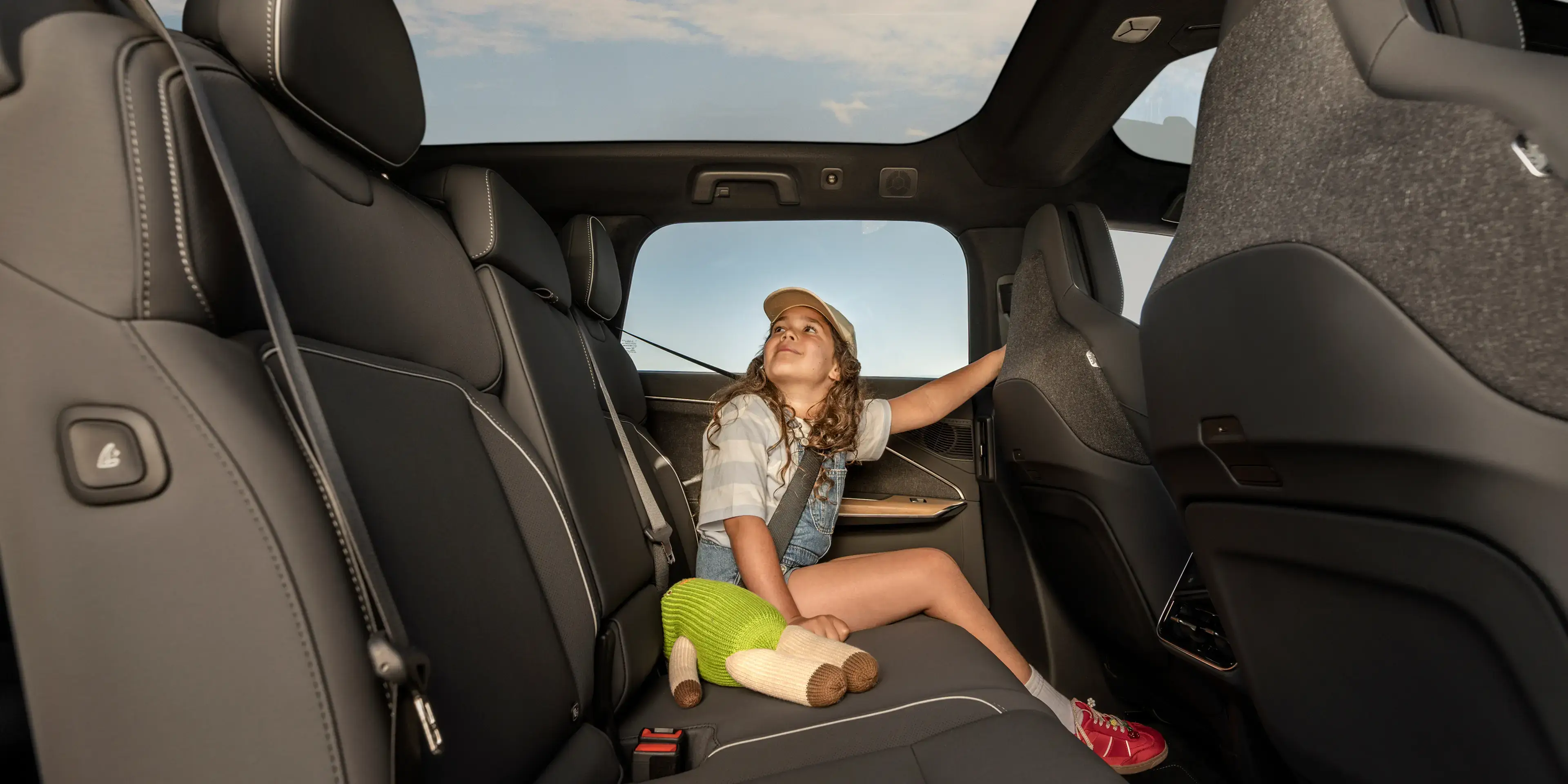 A young girl sits in a dark gray SUV back seat, looking up through a sunroof at a blue sky, holding a stuffed toy.