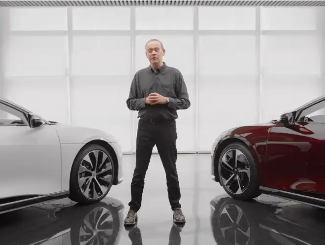 Peter Rawlinson standing between white and red cars in a bright, modern showroom with reflective floor.