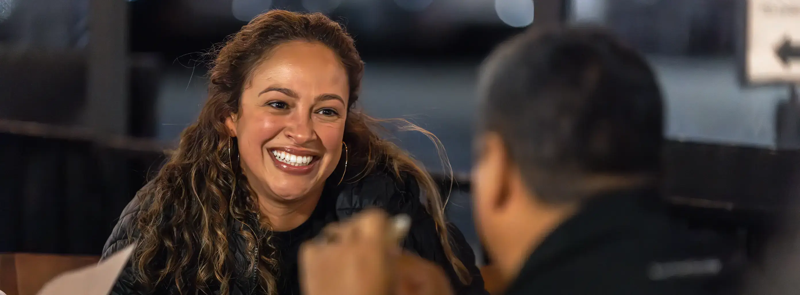 Woman with curly hair in black jacket smiling brightly during conversation at dimly lit restaurant or café.