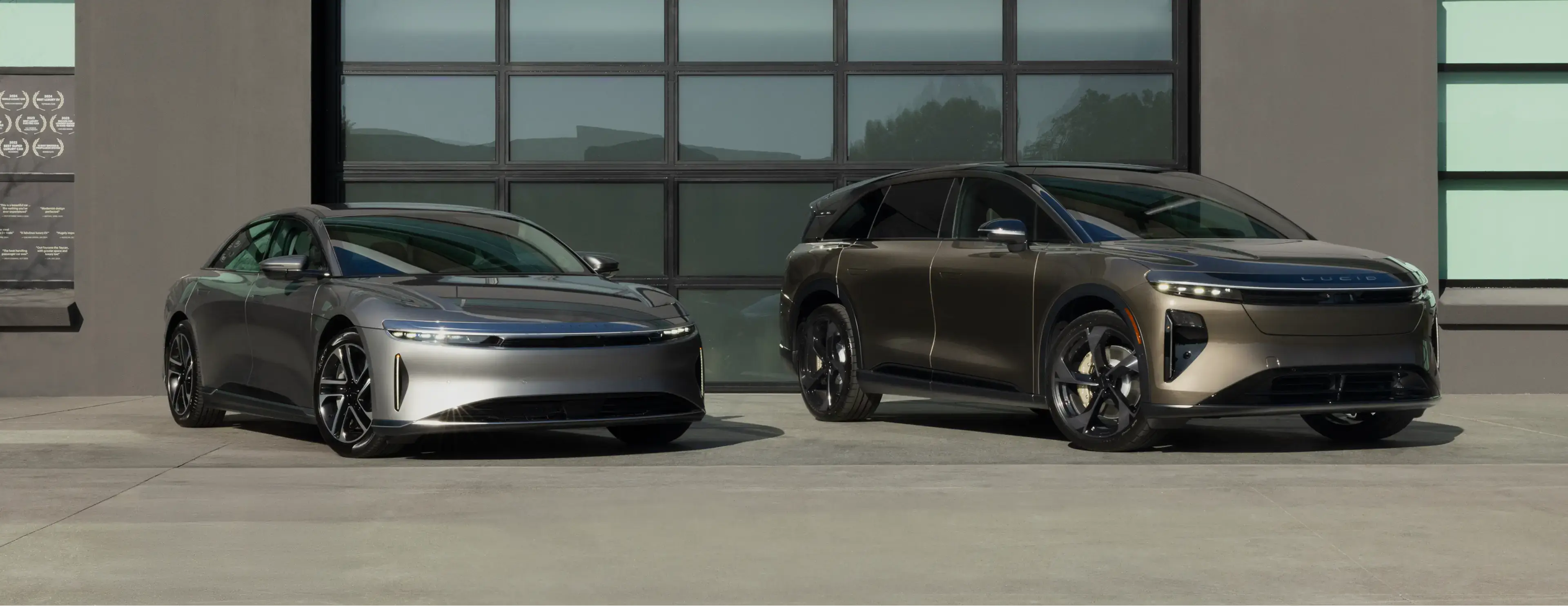 Two sleek electric vehicles parked in front of a modern building with glass windows - a silver sedan and bronze SUV.