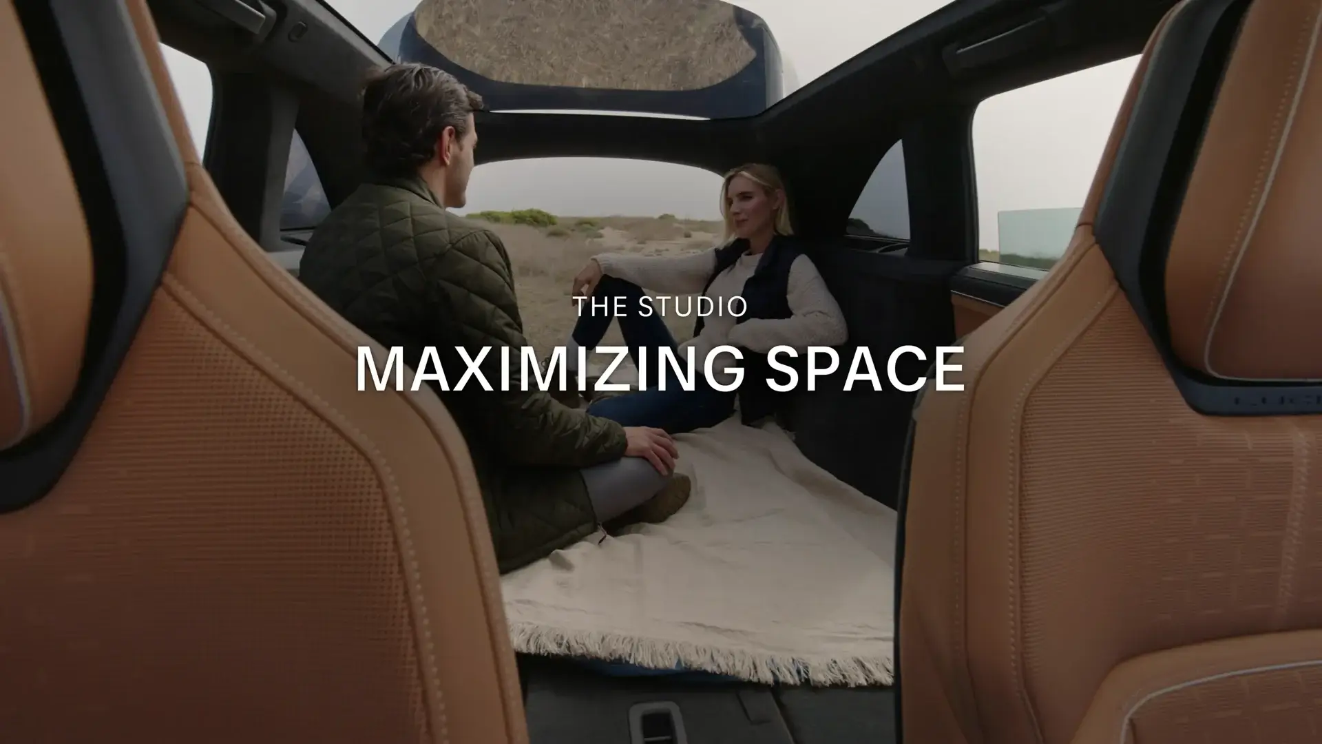 Two people sitting in a spacious vehicle interior with panoramic roof, tan leather seats, and text reading "THE STUDIO MAXIMIZING SPACE"