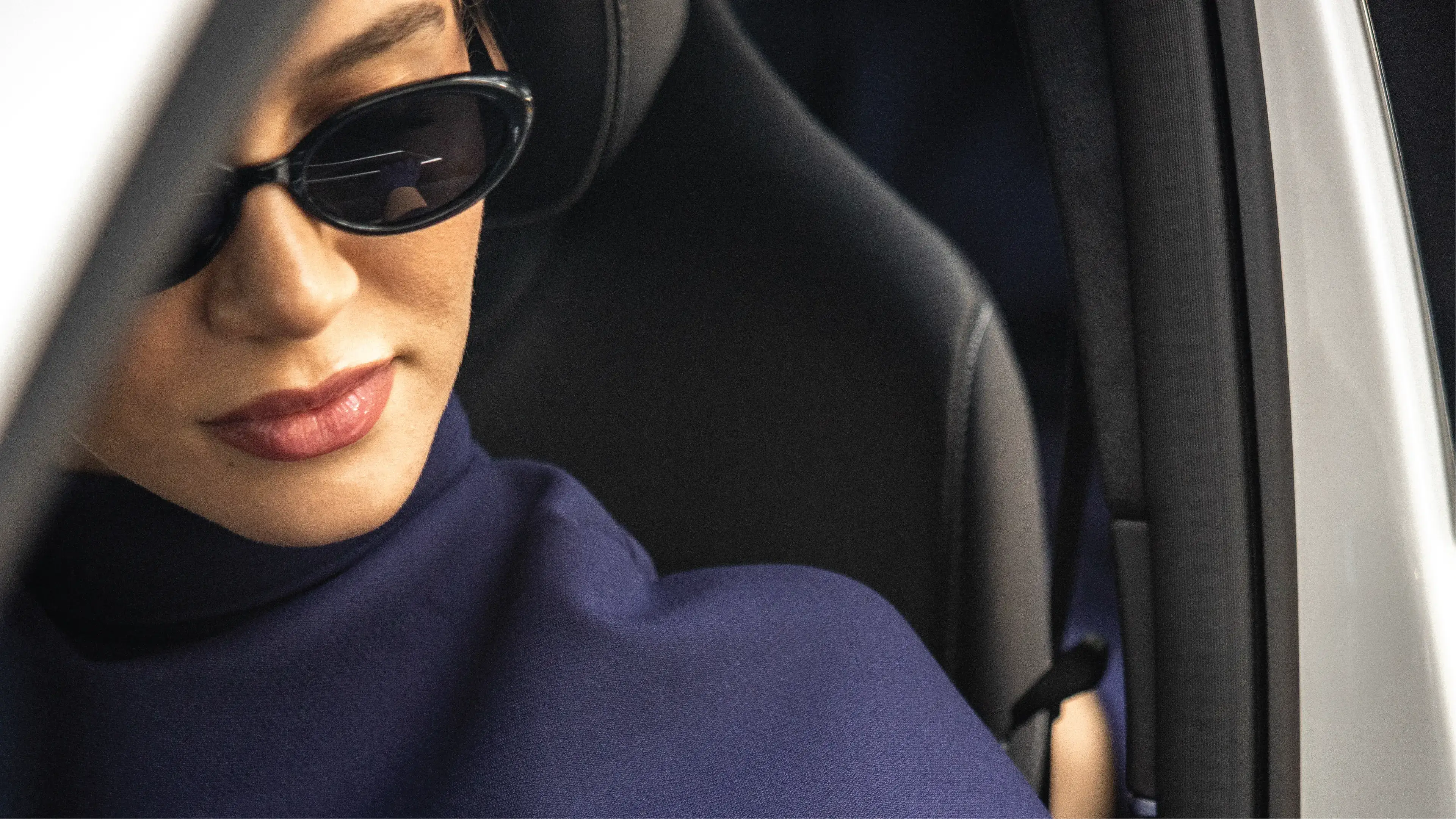 A woman with dark hair and sunglasses, wearing a navy blue turtleneck, is sitting in the passenger seat of a car.
