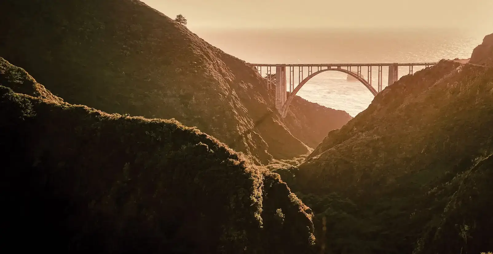 A concrete arch bridge spans rugged coastal cliffs at golden hour, with the ocean glowing softly in the warm background.