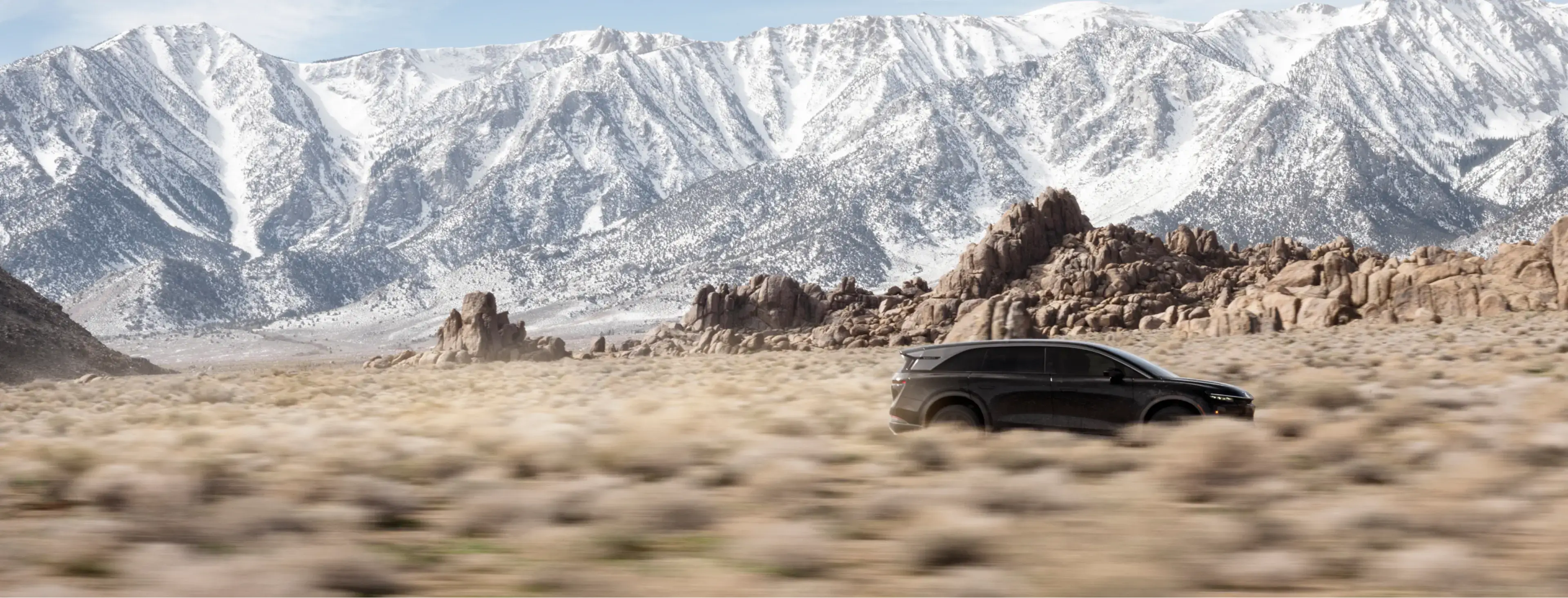 A Lucid Gravity SUV drives through a desert landscape with rocky formations and snow-capped mountains under a blue sky.